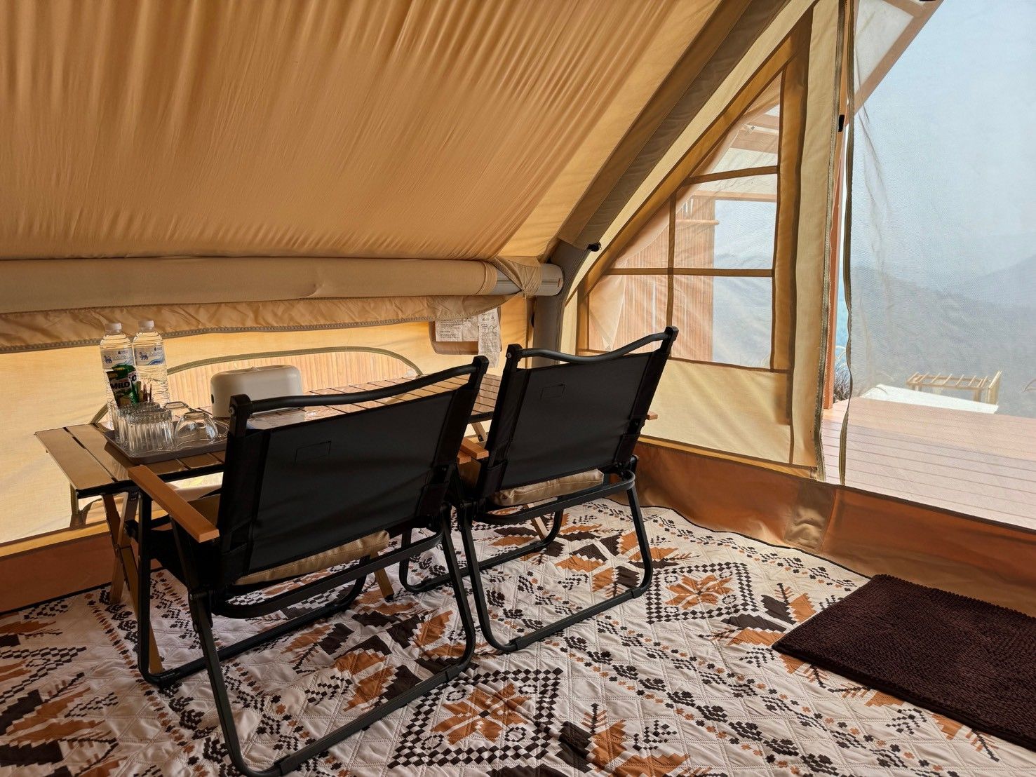 A tented room with a table, two chairs, a patterned floor, and a view through a window.