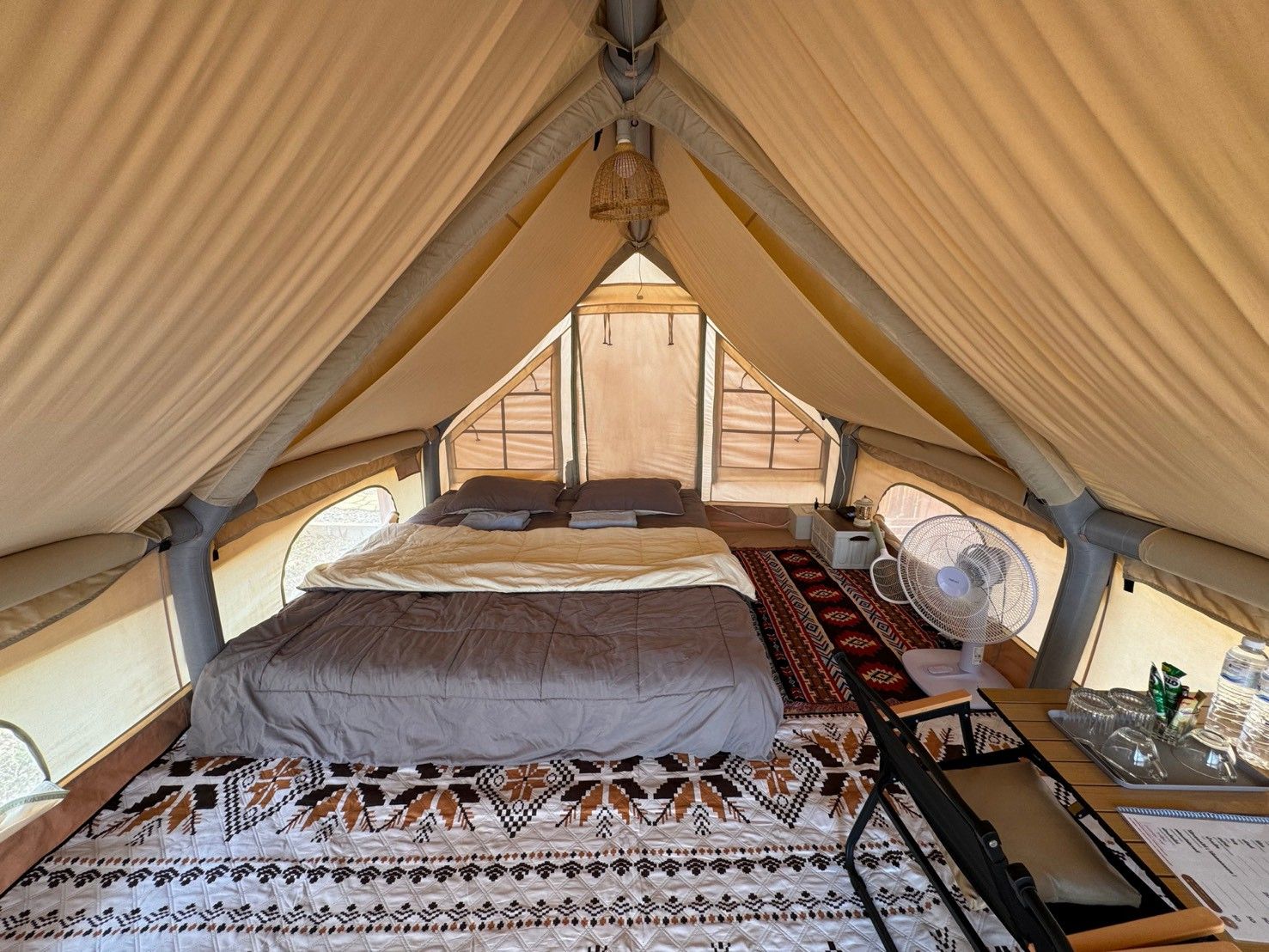 A tent with a bed, a fan, and a table with items.