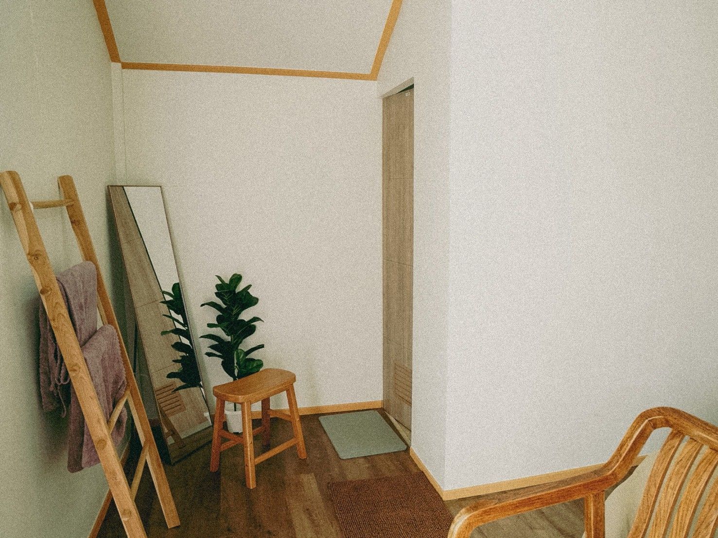 A corner room with a wooden chair, a small stool, a mirror, and a potted plant.