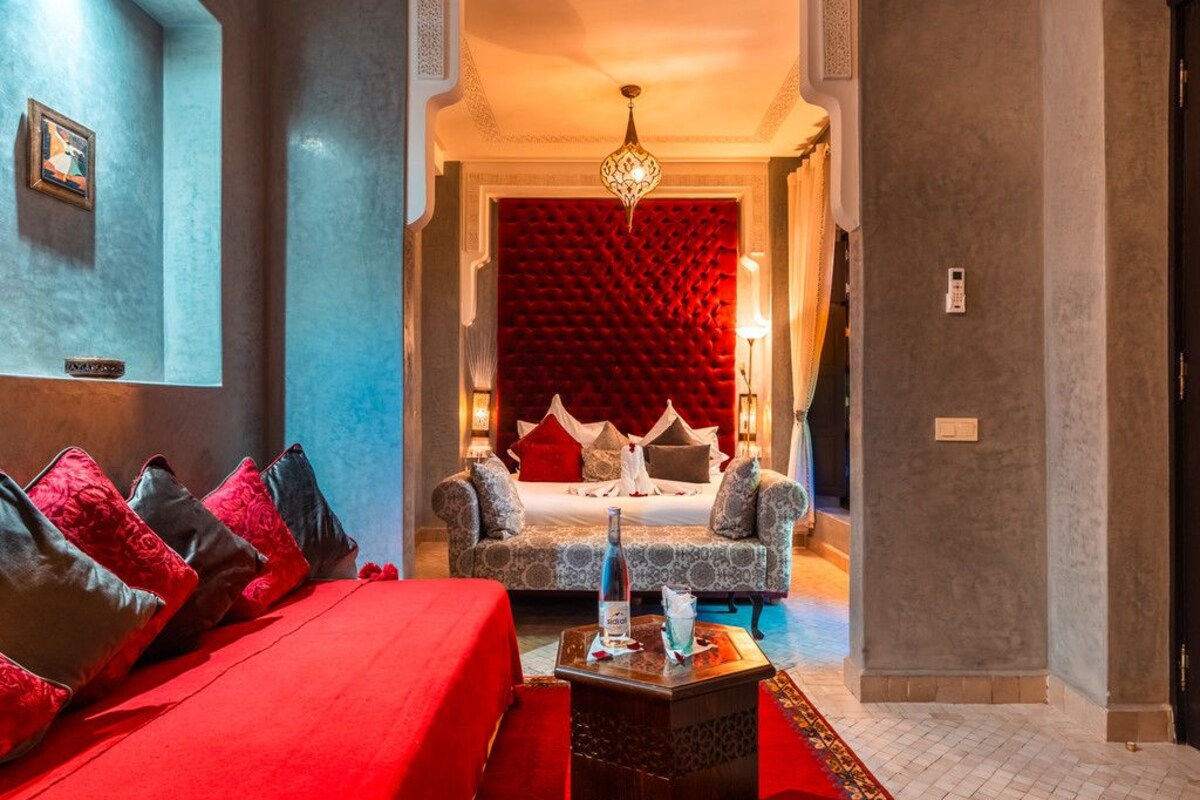 Quarto de hotel com carpete vermelho, cama com roupa de cama branca, mesa de cabeceira com uma garrafa e parede vermelha com padrão texturizado.