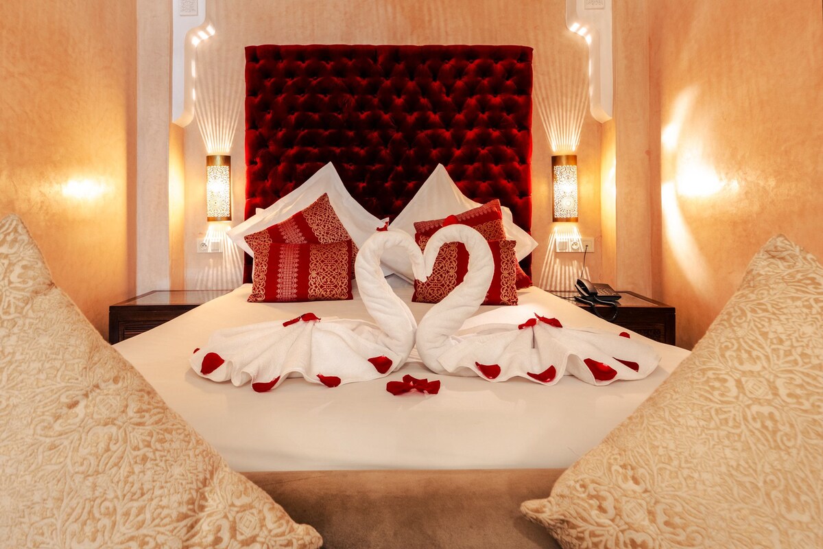 Quarto de hotel com cama, almofadas vermelhas e brancas, cabeceira vermelha e um cisne decorativo feito de toalhas.