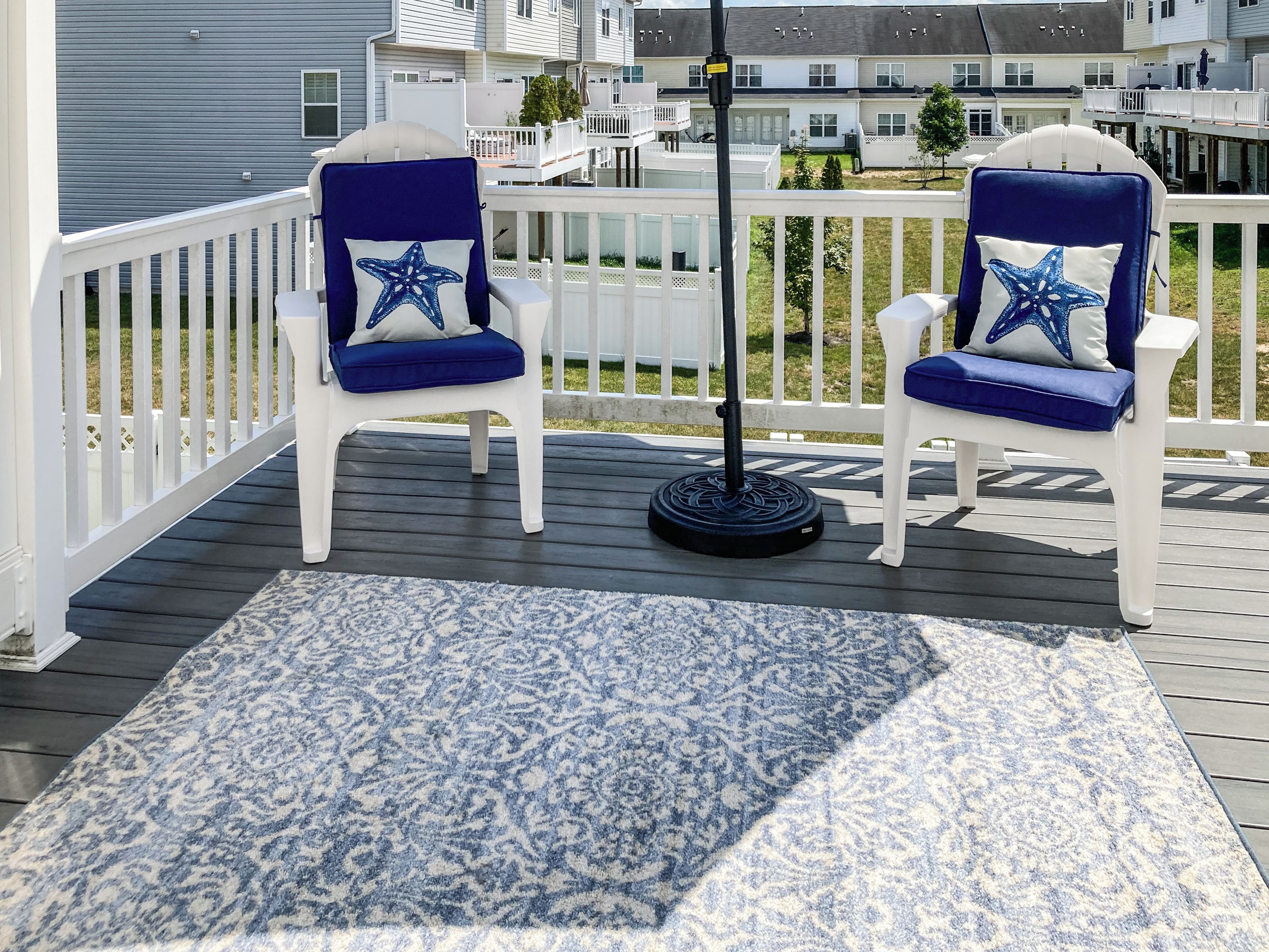 A deck with two white chairs with blue cushions and star-shaped pillows, a patterned rug, and a metal pole.