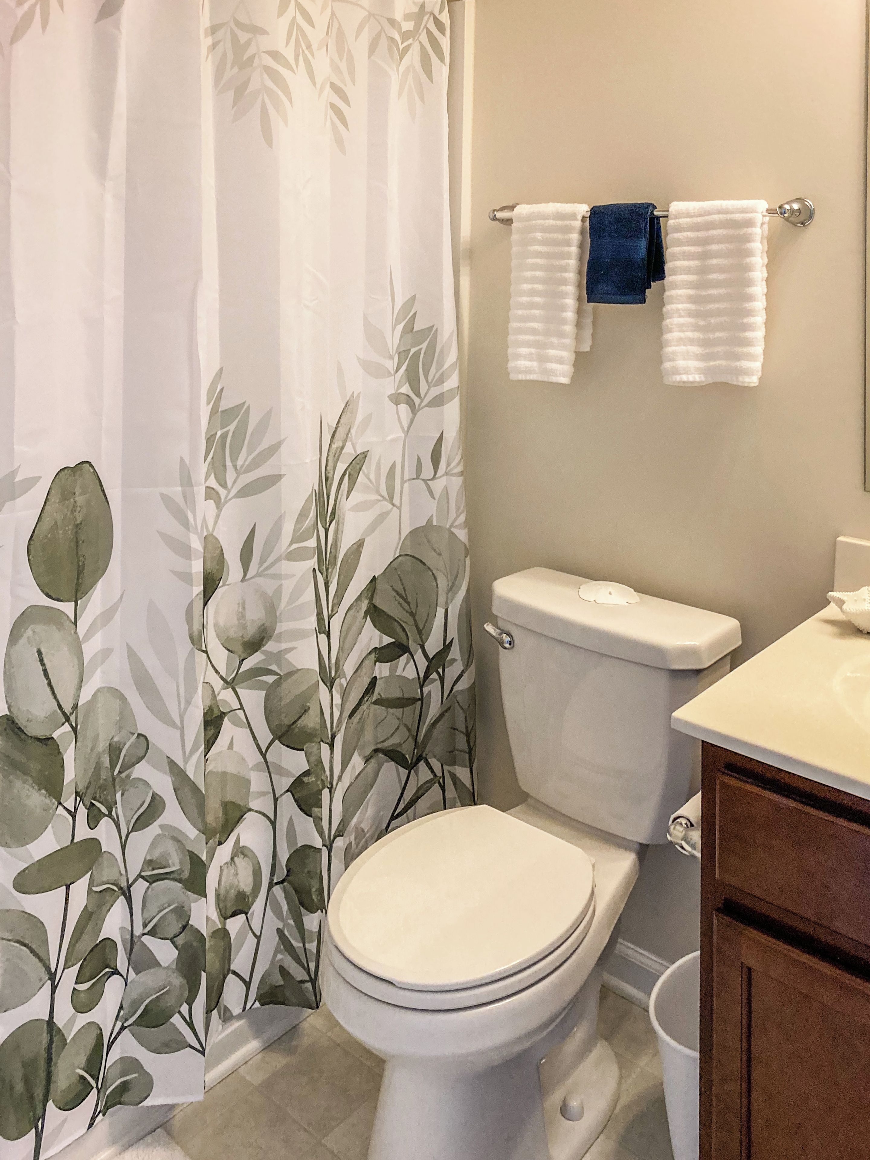 A bathroom with a toilet, a shower curtain with a leaf pattern, and a towel rack with two towels.