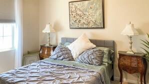 Une chambre à coucher avec un lit, deux tables de chevet, un tableau et une fenêtre avec des stores.