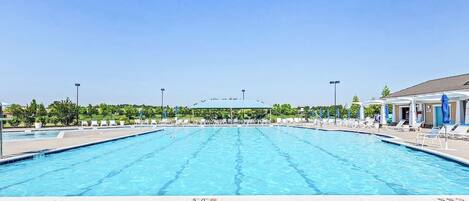 Una amplia piscina al aire libre con múltiples carriles, rodeada de sillas de descanso y sombrillas, y un área cubierta al fondo.