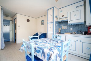 Une cuisine compacte avec un coin repas, comprenant une table recouverte d’une nappe bleue et blanche, une chaise bleue et un plan de travail avec un évier et une cuisinière.