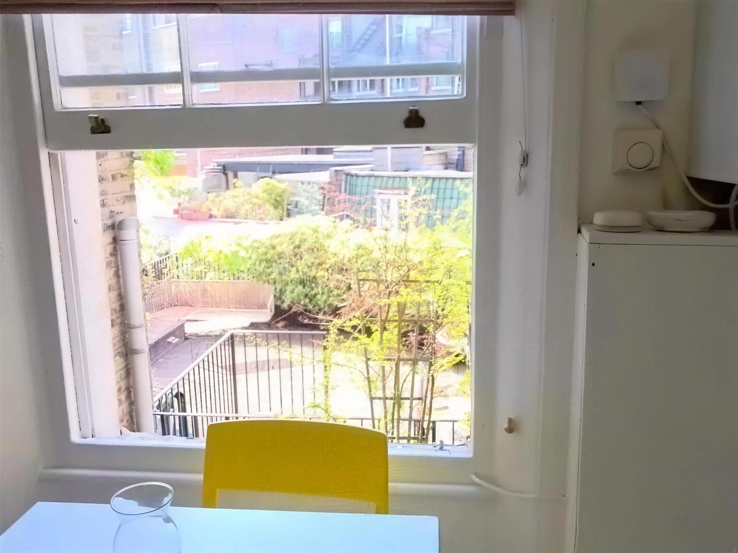 Una habitación con ventana que da a un jardín, una mesa blanca y una silla amarilla.