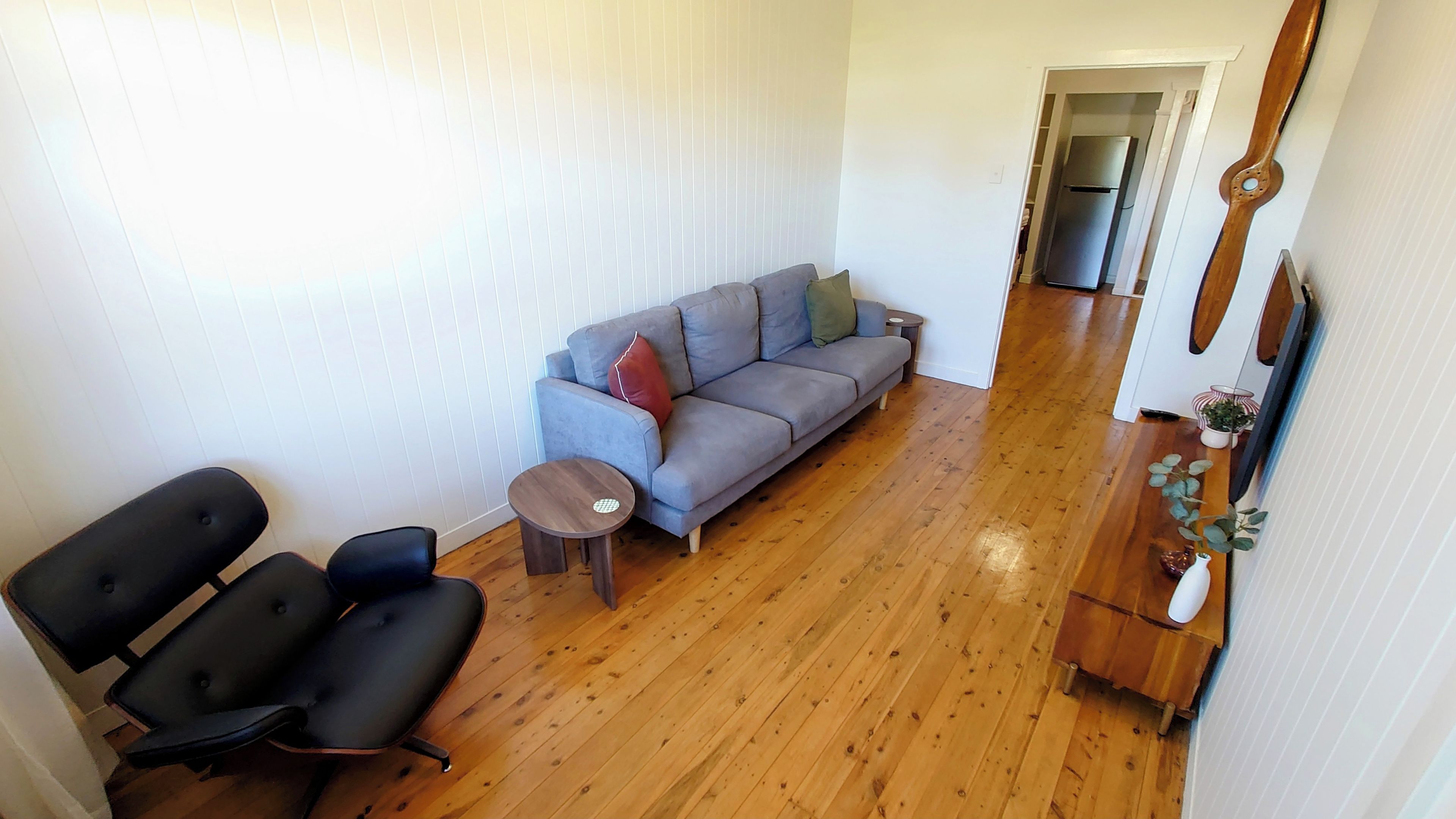 A living room with a grey sofa, two black chairs, a small round table, wooden flooring, and a doorway leading to a kitchen.