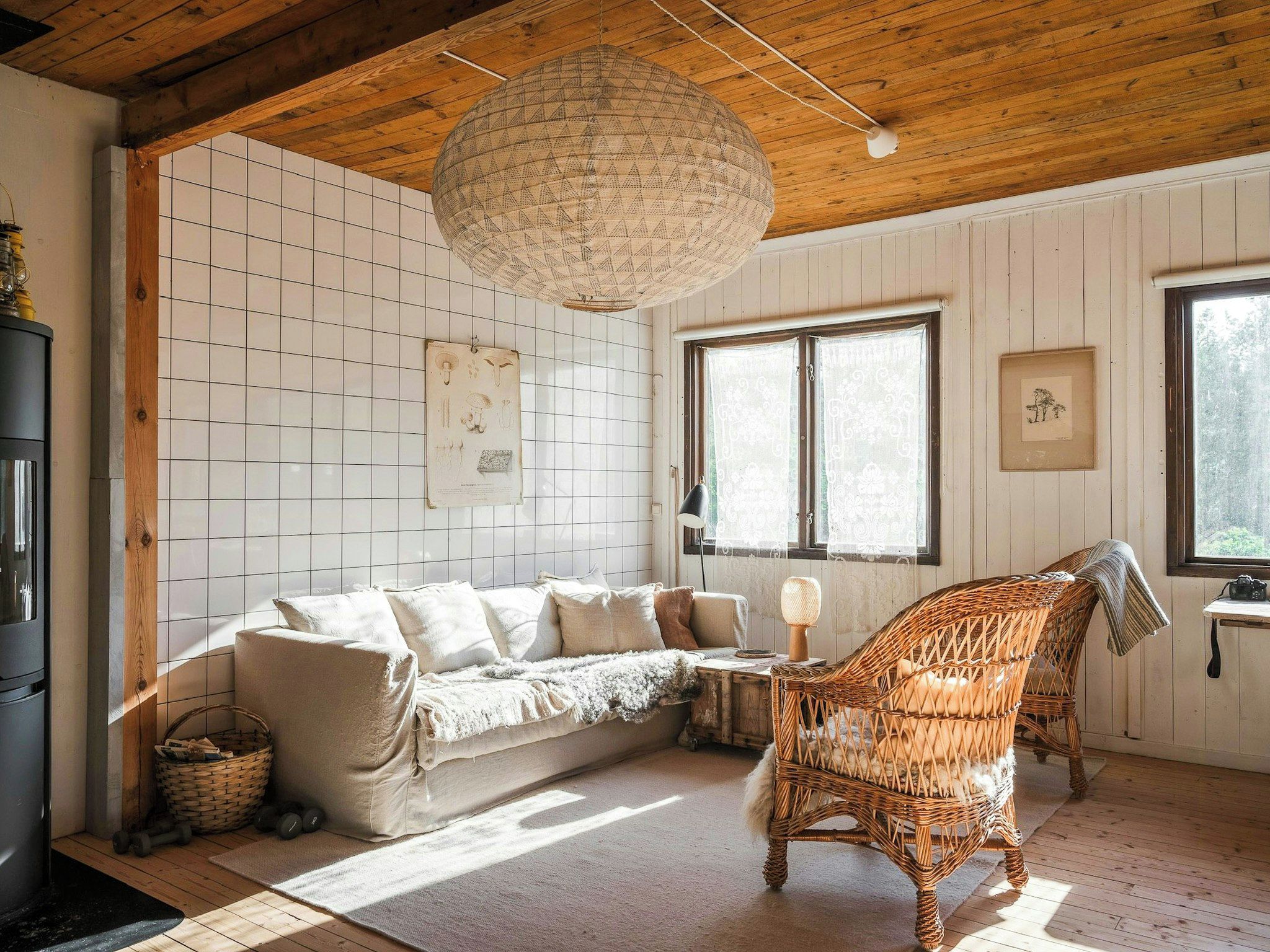 Une pièce chaleureuse avec un plafond en bois, un luminaire tressé, un canapé, un fauteuil en osier et une fenêtre ornée de rideaux à motifs.