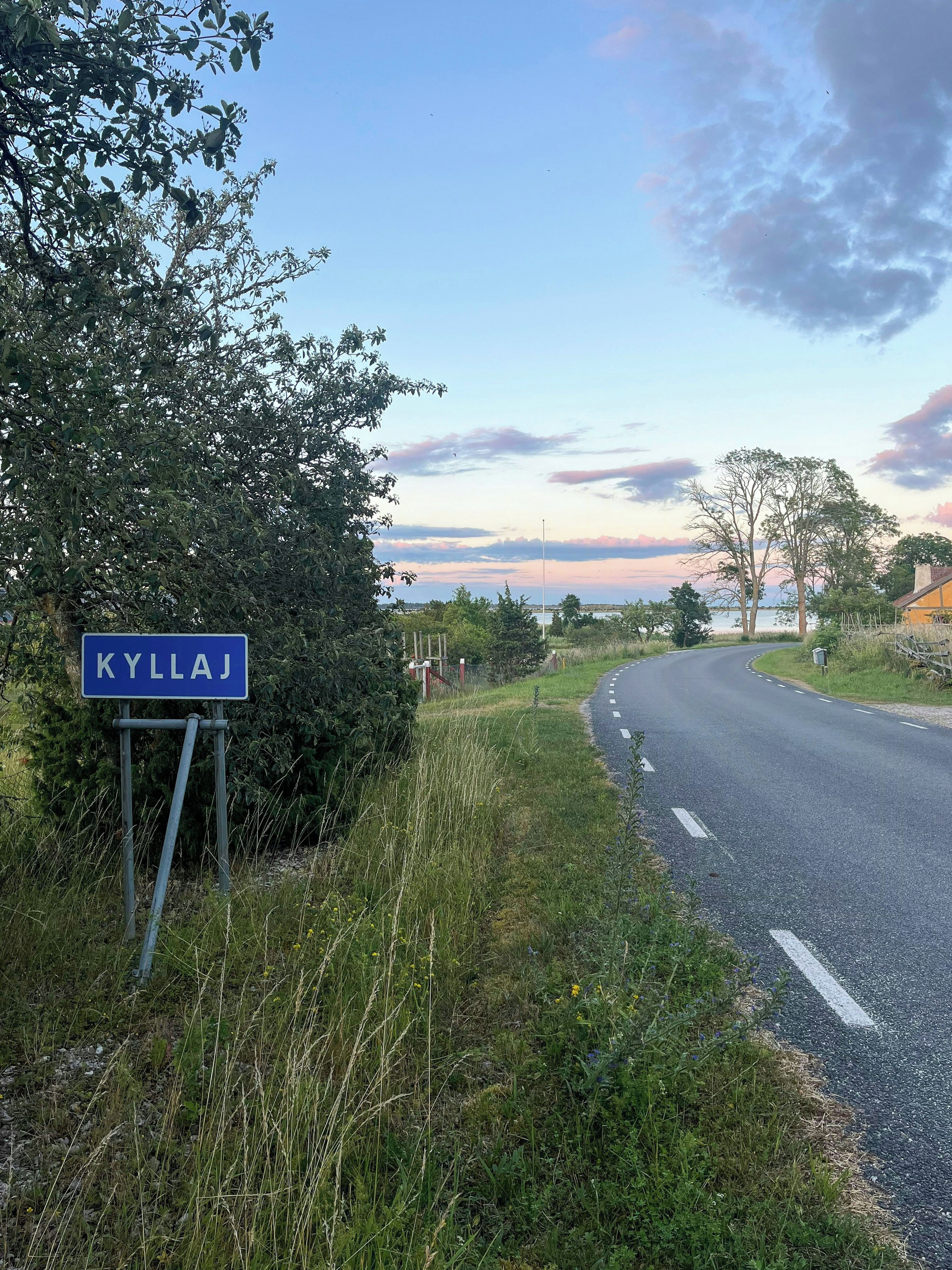 Een wegwijzer met de naam 'KYLLAJ' en een landelijk landschap met bomen en een heldere lucht.