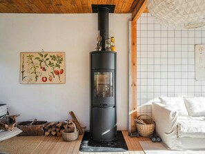 Ein Zimmer mit einem schwarzen Kaminofen, einer weiß gefliesten Wand, einer Holzbalkendecke, einer Katze, einem Holzscheitkorb und einem weißen Sofa.
