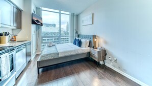 A bedroom with a bed, a nightstand, a TV, a window with a city view, and a kitchenette. - StayToronto - CityLite Studio Stay in Toronto - DT (Toronto)