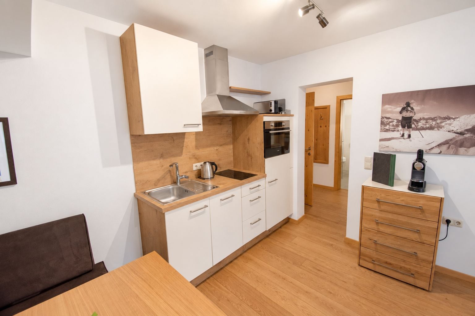 Een compacte keuken met een spoelbak, fornuis en keukenkastjes. Er is een eethoek met een tafel en stoelen.
