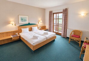 Habitación de hotel con una cama grande, dos mesitas de noche, una silla, un ventanal con cortinas y una lámpara de pared.