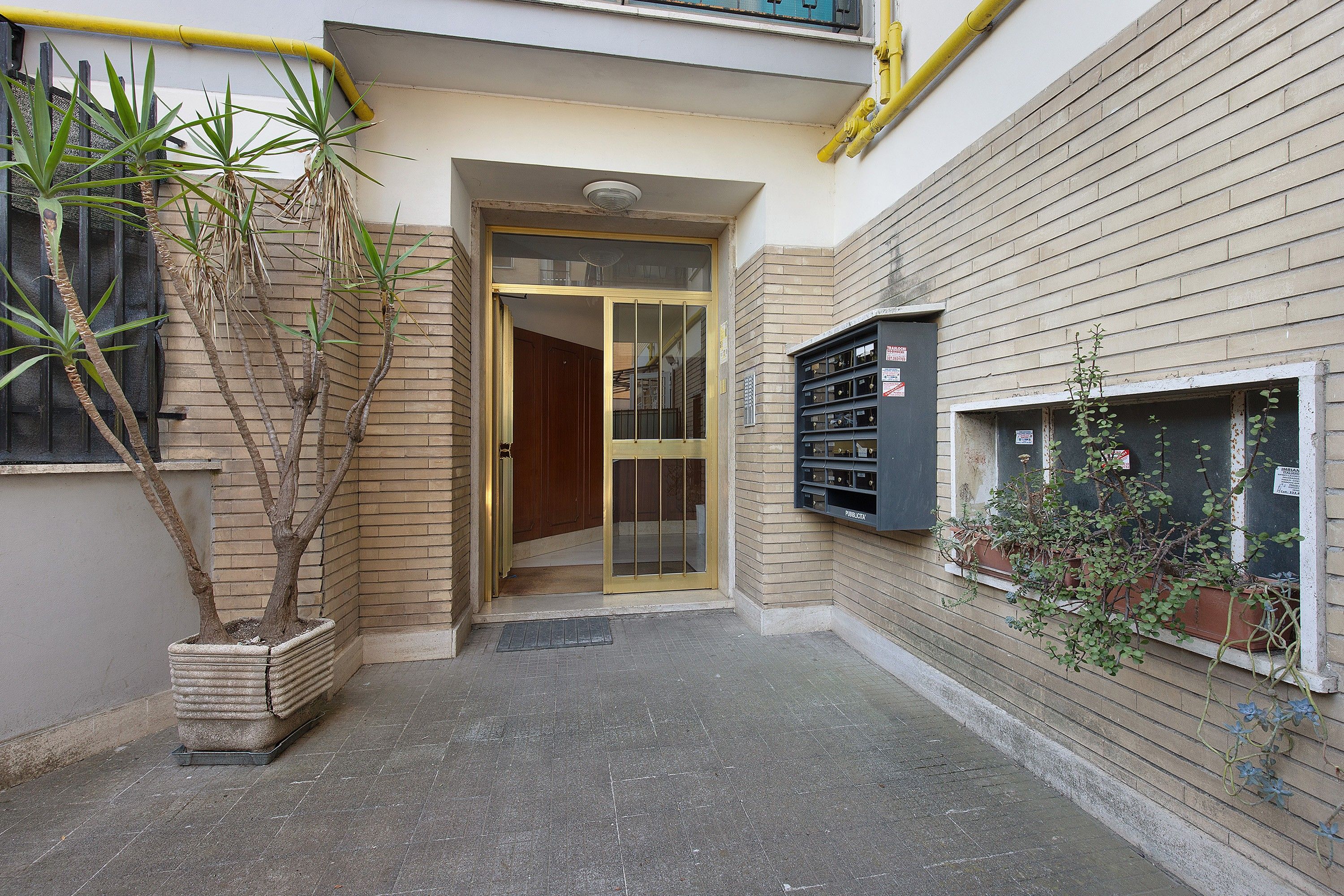 Entrada de un edificio con puerta de vidrio, buzón y plantas en macetas.