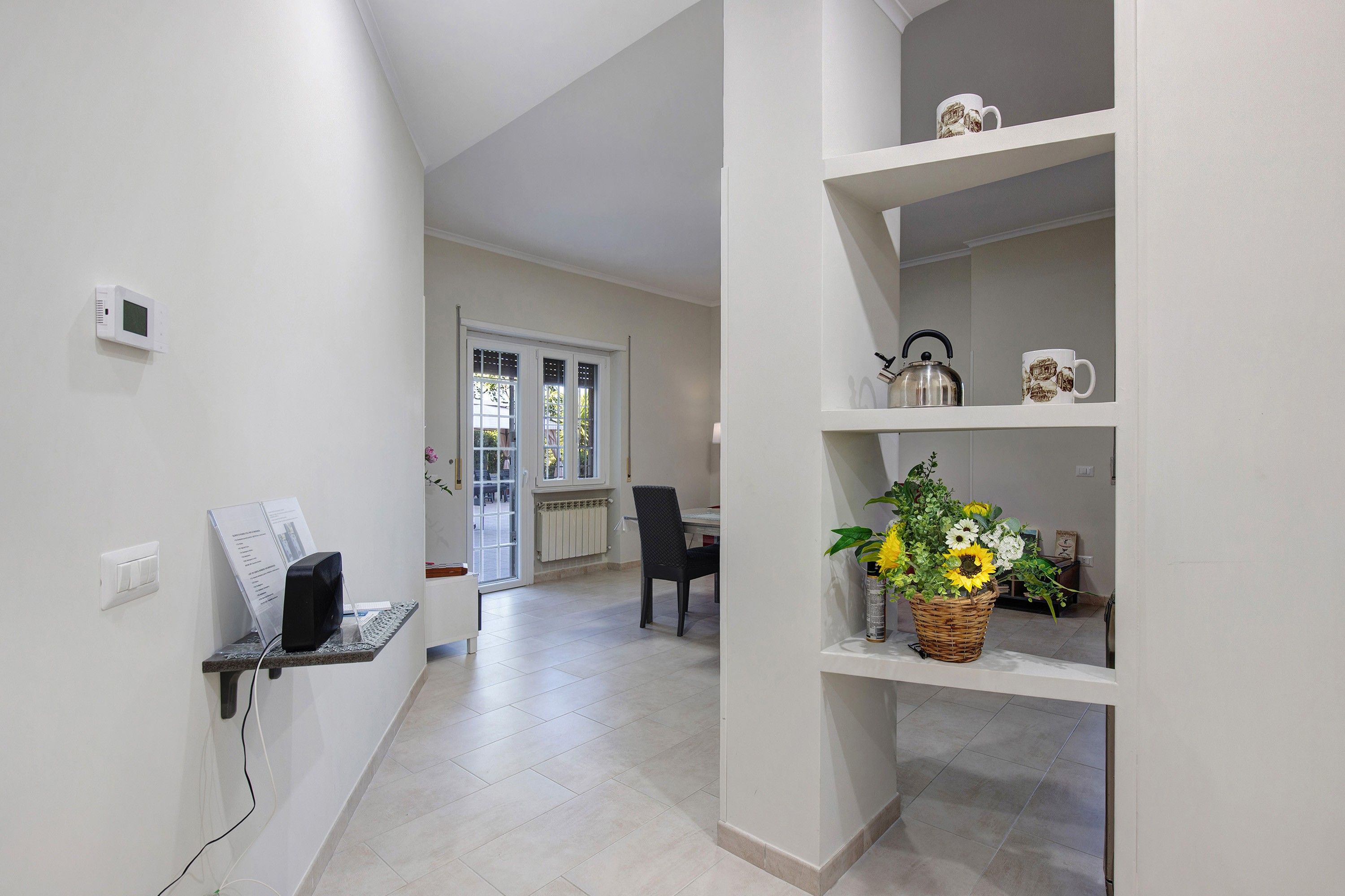 A hallway with a white wall, a thermostat, a power outlet, and a shelf with a kettle and a mug.