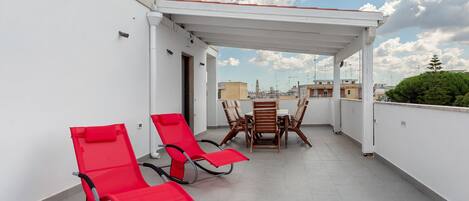 A rooftop terrace with two red lounge chairs, a wooden table, and a white pergola.