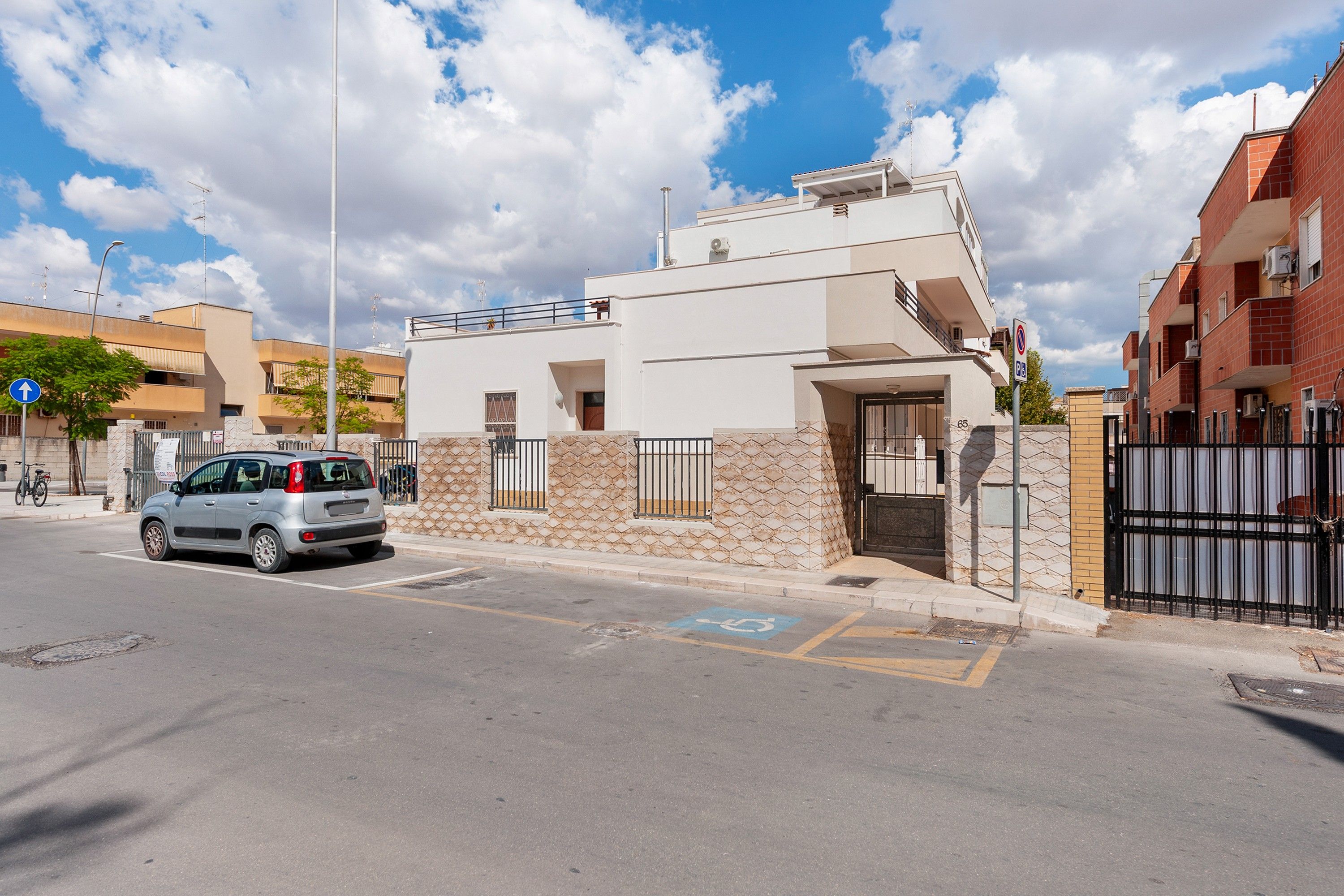 Un edificio moderno con fachada blanca, entrada protegida por una cancel y una plaza de estacionamiento señalizada con el símbolo de una silla de ruedas.