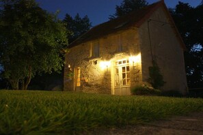 Ein Steinhaus mit geneigtem Dach, beleuchtet durch Außenlichter, umgeben von einer Rasenfläche und Bäumen.