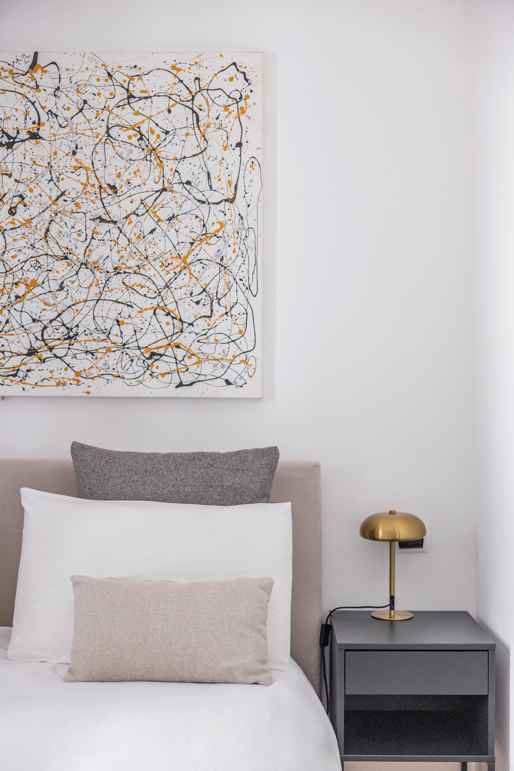 Un dormitorio con cama, almohadas, una mesita de noche, lámpara y un cuadro abstracto de gran tamaño en la pared.