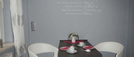 A dining area with a table set for two, a wall with a decorative phrase, and a white chair.