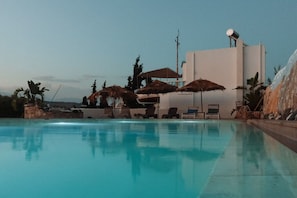 Une piscine avec des chaises longues et des parasols, un bâtiment équipé d’une antenne parabolique et un ciel dégagé.