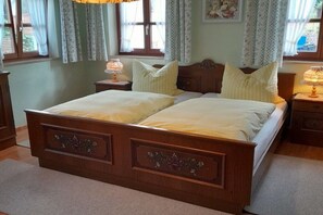 Ein Doppelbett mit hölzernem Gestell und floralen Verzierungen, begleitet von Nachttischen mit Lampen, in einem Zimmer mit Fenster und Vorhängen.