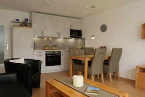 A modern kitchen with a dining area, including a table with chairs, a sofa, and a clock on the wall. - Holiday home with 1 bedroom (Wurster Nordseeküste)