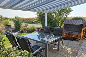 An outdoor patio with a glass table, four chairs, a barbecue grill, and a covered seating area. - Holiday home with 1 bedroom (Wurster Nordseeküste)