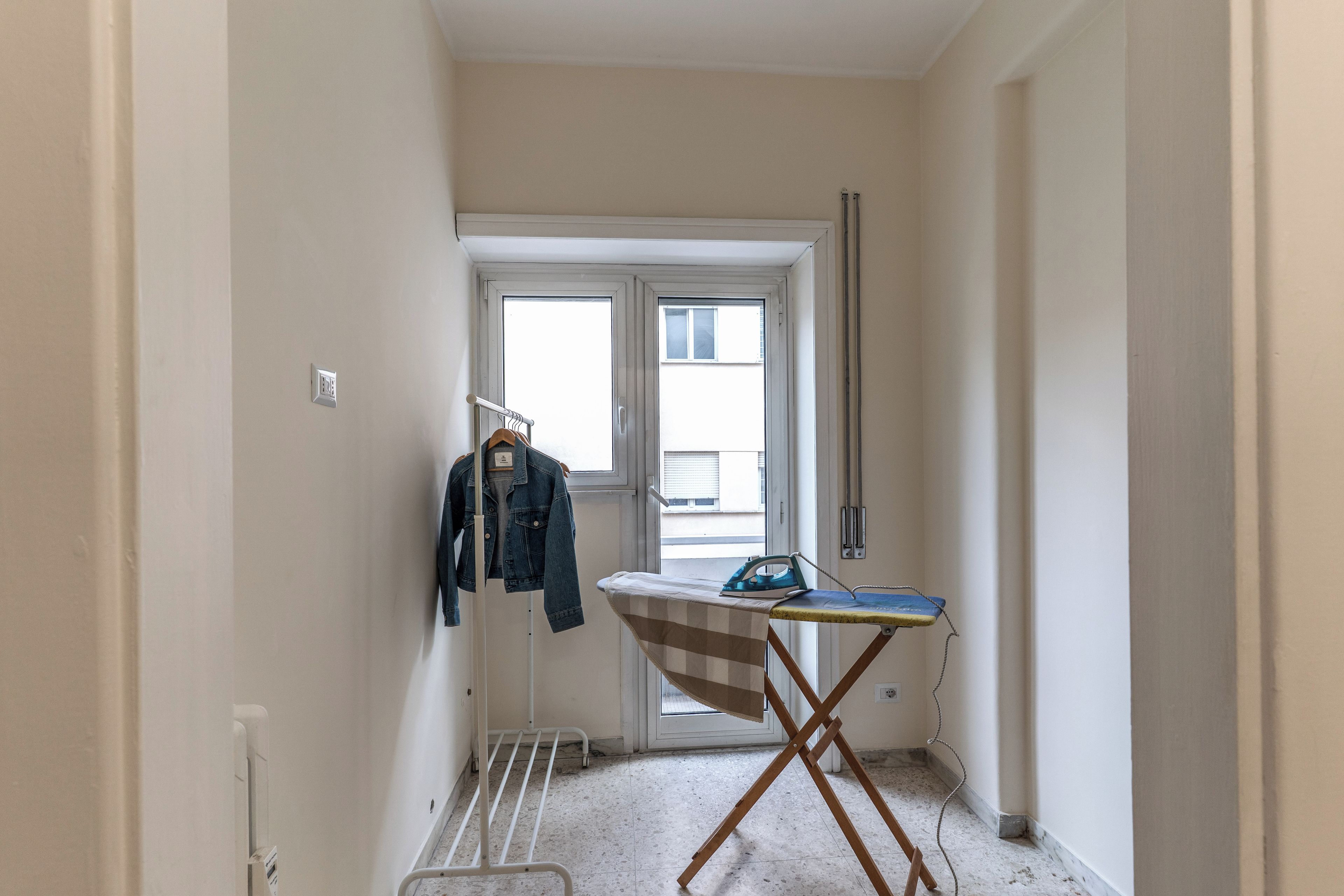 Een smalle gang met een deur naar een kamer. Een kledingrek hangt er en er staat een strijkplank met een stoomstrijkijzer in de kamer.