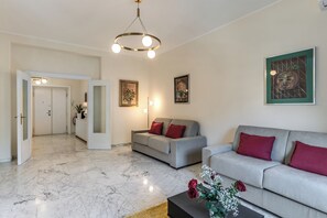 Un salon moderne avec un canapé gris, une table basse et un sol en marbre. Il y a deux portes qui donnent sur d’autres pièces, et un tableau accroché au mur.