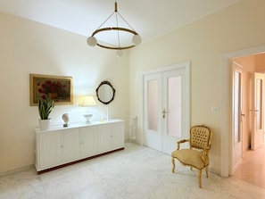 A room with a white cabinet, a painting of flowers, a mirror, a door with a window, and a chair.