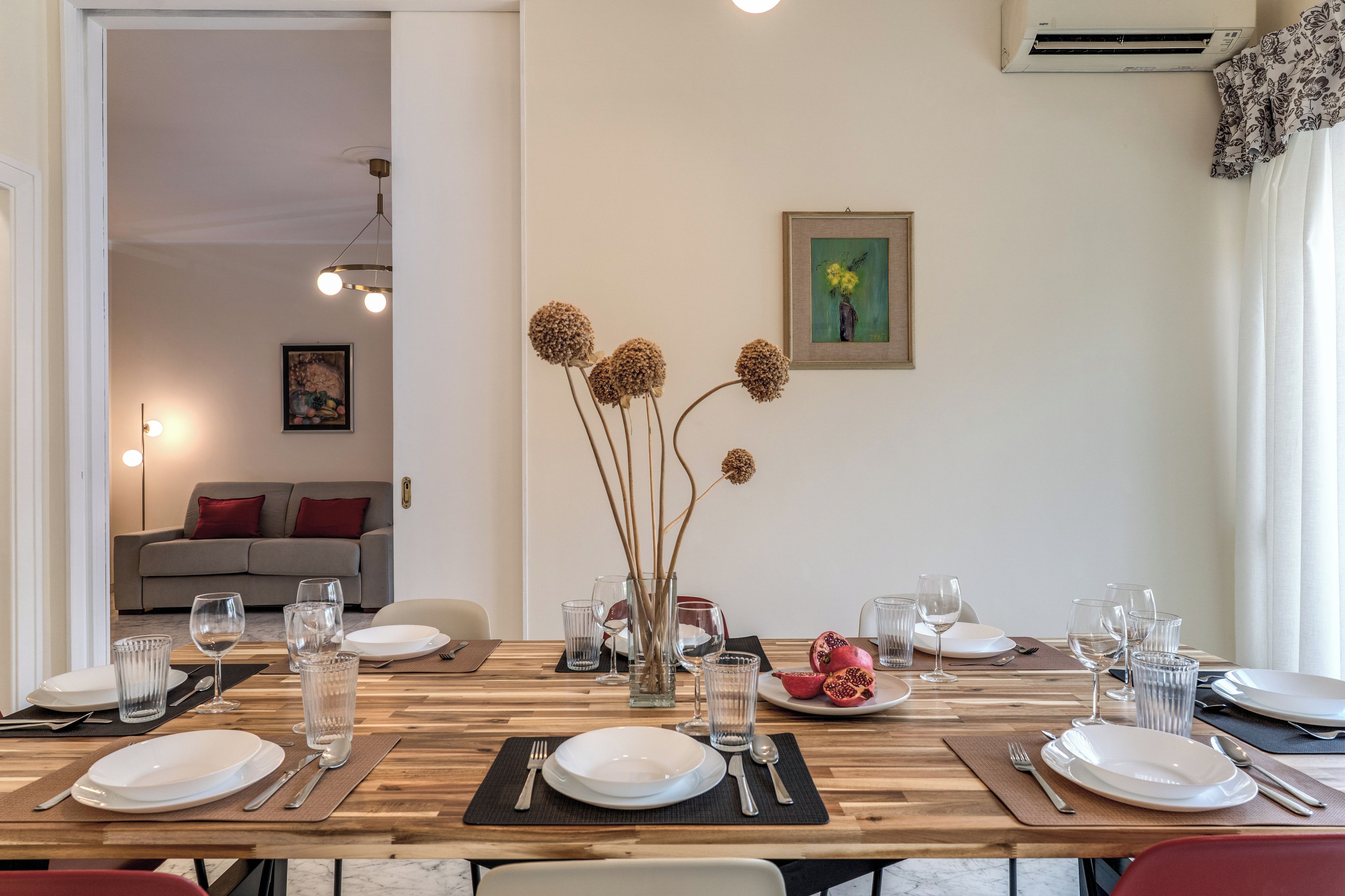 Zona pranzo con tavolo in legno apparecchiato per un pasto, un vaso con fiori secchi e un quadro appeso al muro.