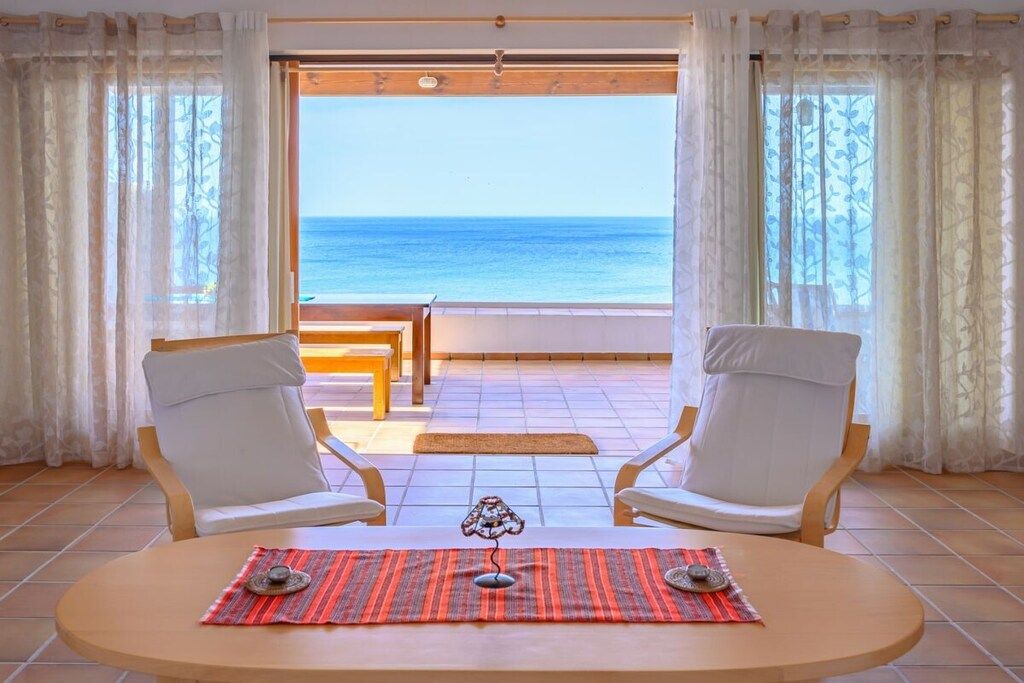 A room with a view of the sea, two white chairs, a table with a placemat, and a small lamp.
