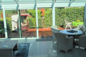 A conservatory with wicker furniture, a glass table, and a view of a garden. - Holiday home with 2 bedrooms (Wangerland)