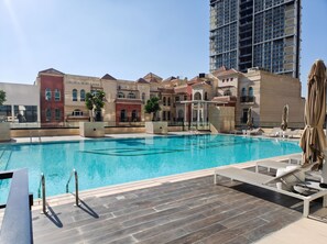 A swimming pool with lounge chairs and umbrellas, surrounded by multi-story buildings with balconies. - StayShort - Peace and Tranquility in this 1 Bedroom in JVC (Dubai)