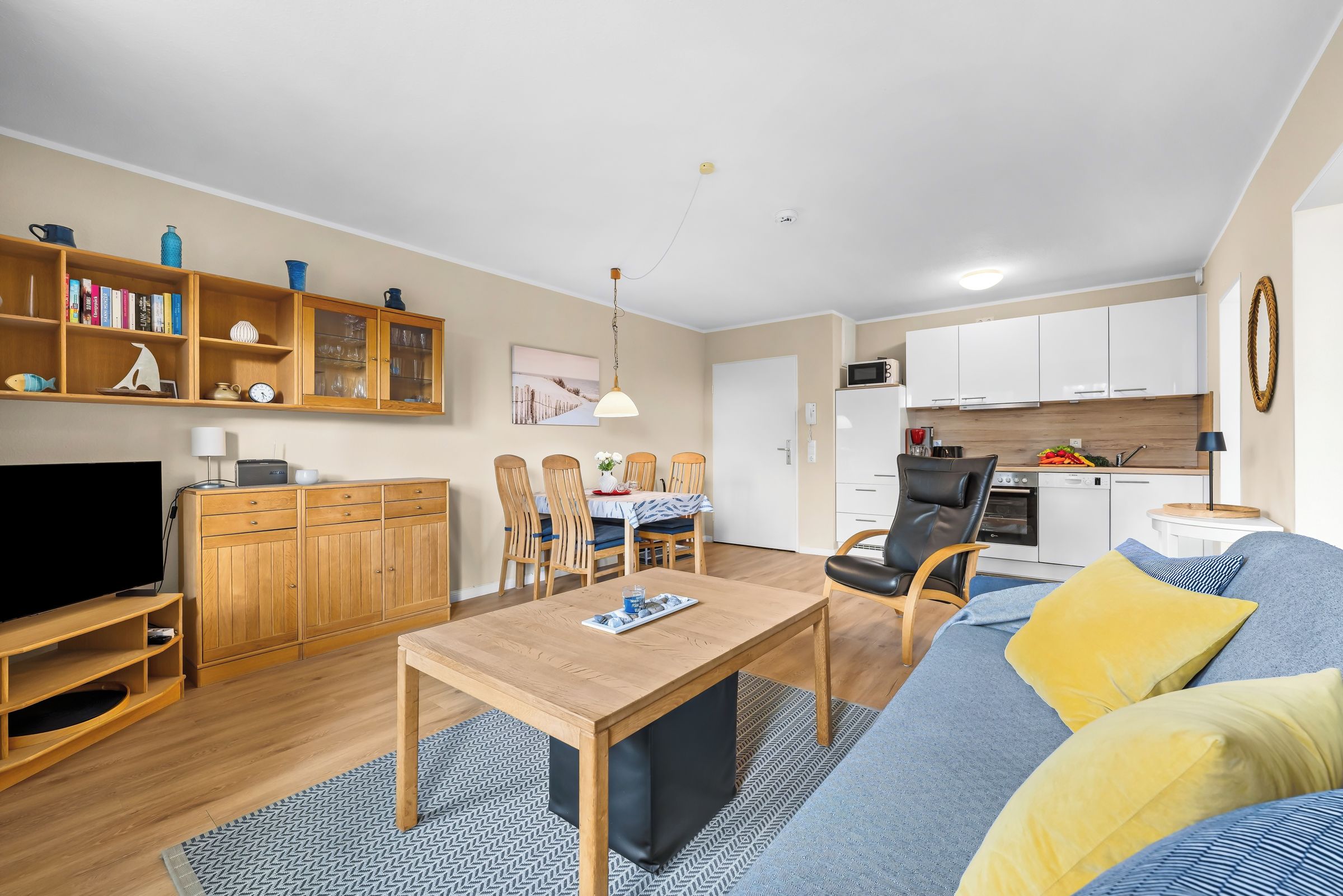 A modern living room with a flat-screen TV, wooden furniture, a dining area with chairs, and a kitchen with white cabinets.