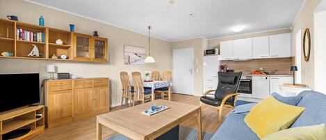 A modern living room with a flat-screen TV, wooden furniture, a dining area with chairs, and a kitchen with white cabinets.