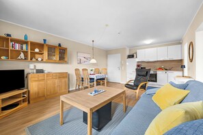 Ein modernes Wohnzimmer mit Flachbildfernseher, Holzmöbeln, einem Essbereich mit Stühlen und einer Küche mit weißen Schränken.