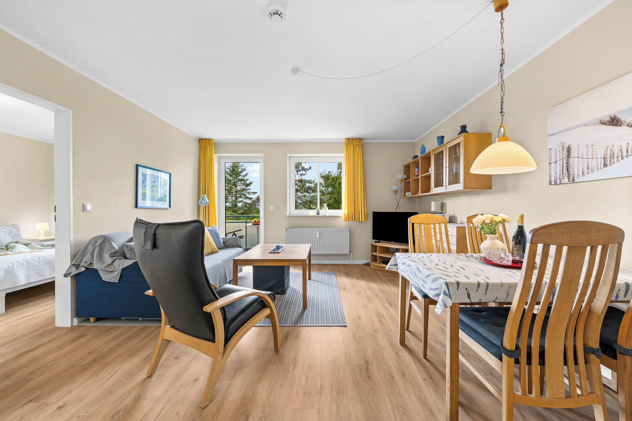 A well-lit, open-plan living area with a sofa, dining table, and chairs, a kitchen with a TV, and large windows with curtains.