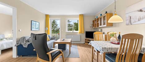 Un espacio habitable amplio y bien iluminado, con sofá, mesa de comedor y sillas, cocina con televisor y ventanas grandes con cortinas.