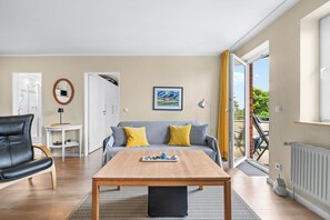 Ein modernes Wohnzimmer mit einer Couch, einem Couchtisch und einem Esstisch. Eine Tür führt zu einem Badezimmer, ein Fenster mit Vorhängen und ein Heizkörper sind ebenfalls vorhanden.