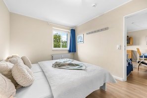 Ein Schlafzimmer mit einem Bett, einem Fenster, einer Heizung, einer Tür zu einem anderen Raum und einem an der Wand befestigten Regal.