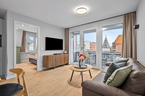 Ein modernes Wohnzimmer mit einer Couch, einem Fernseher, einem Holzschrank und einem Balkon mit Blick auf Gebäude.