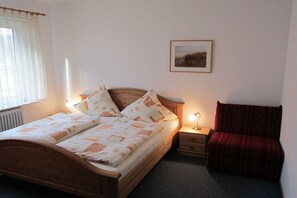 A bedroom with a wooden bed, bedside tables, a red chair, a window with curtains, and a framed picture on the wall. - Apartment 1 - Seestein Holiday Home (Borkum)