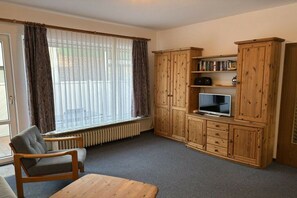 A living room with a sofa, a wooden cabinet with a television, and a window with curtains. - Apartment 1 - Seestein Holiday Home (Borkum)