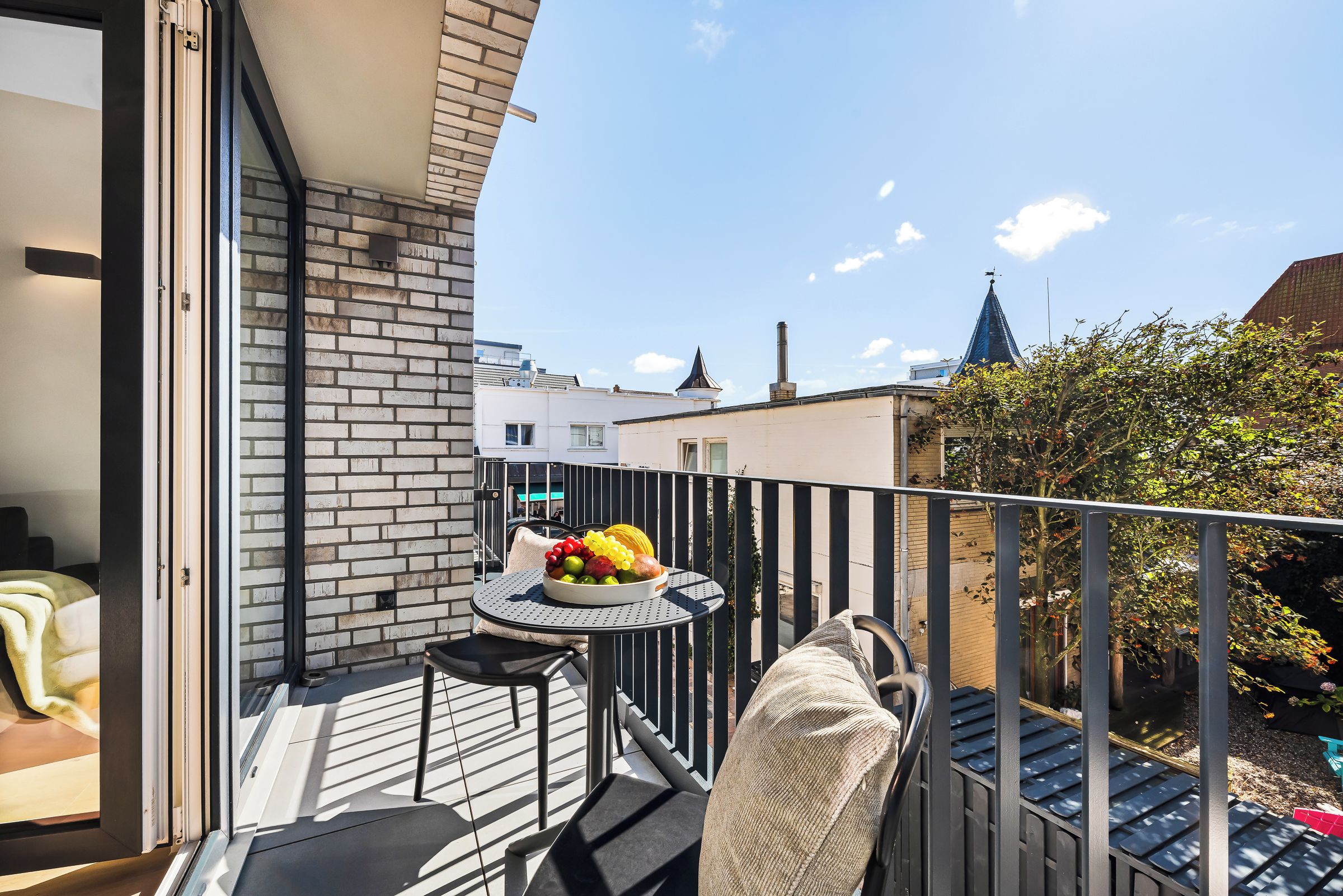 Un balcon avec une table où sont disposés des fruits, une chaise et une vue sur le paysage urbain.
