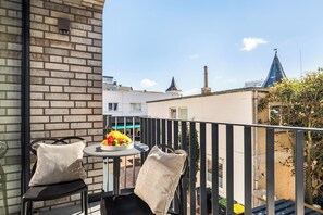 Ein Balkon mit zwei Stühlen, einem kleinen runden Tisch mit Obst und Blick auf Gebäude sowie einen Kirchturm.