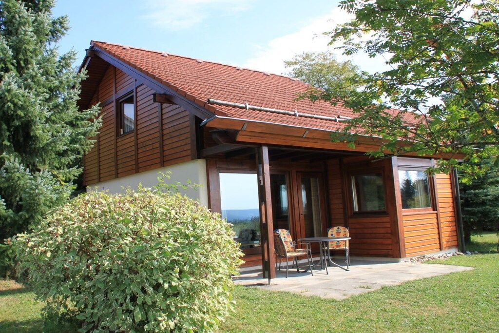 A wooden cabin with a sloped roof, a covered patio with a table and chairs, and a view of the landscape.