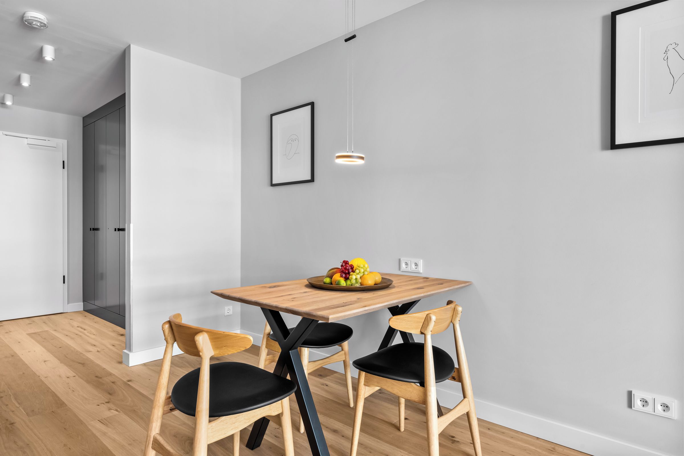 Een moderne eetruimte met een houten tafel en twee stoelen, een fruitschaal op tafel, en een hanglamp erboven.